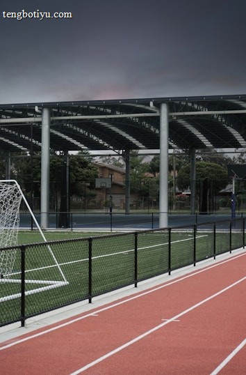 An outdoor sports facility featuring a running track and a soccer goal. The track is red, surrounded by a black metal fence, and the field is covered with artificial green turf. In the background, a basketball court is visible under a covered roof with metal supports. The sky is overcast, adding a moody atmosphere.